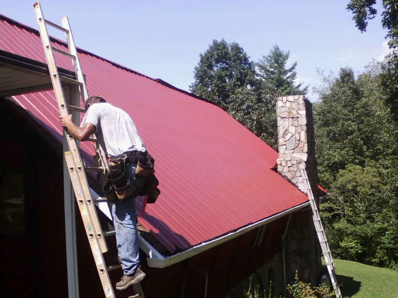 Roofer on a ladder beside a red metal roof and stone chimney — Soffit & Fascia Repair by Roofing Brothers in Ohiopyle